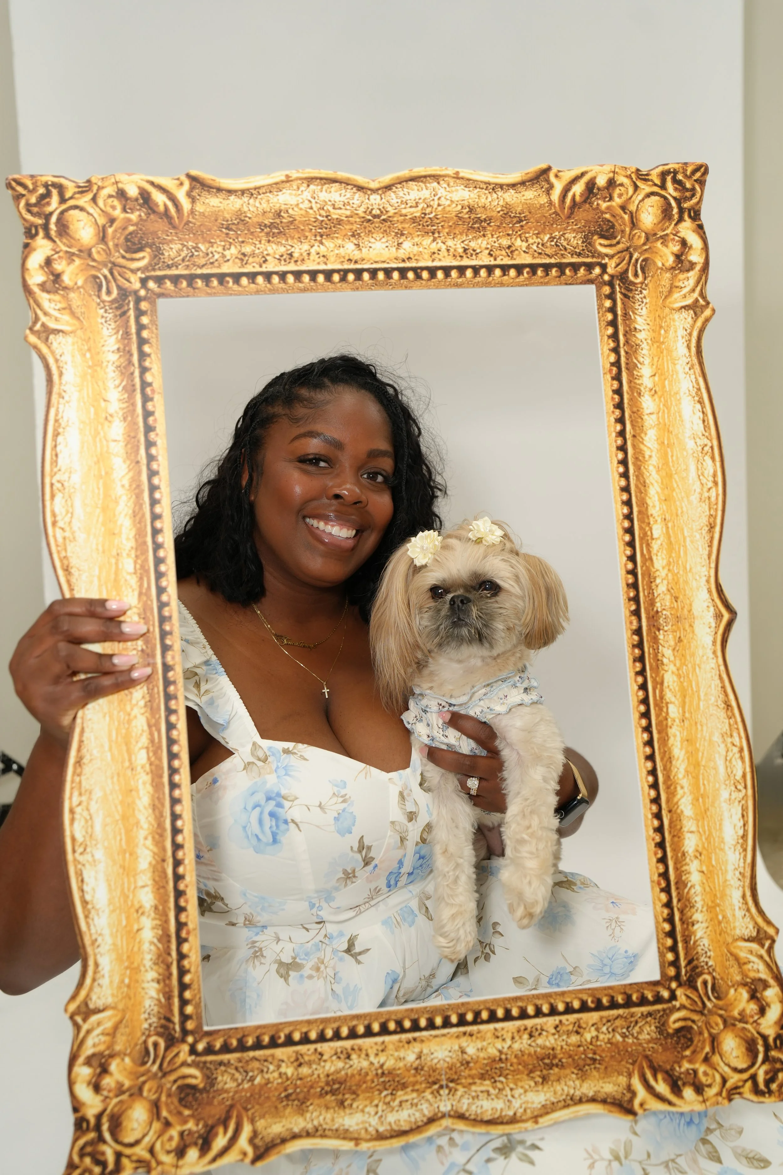 A woman smiling and holding a small dog with flowers in its hair, seen through an ornate gold frame. The woman is wearing a white dress with blue floral patterns.