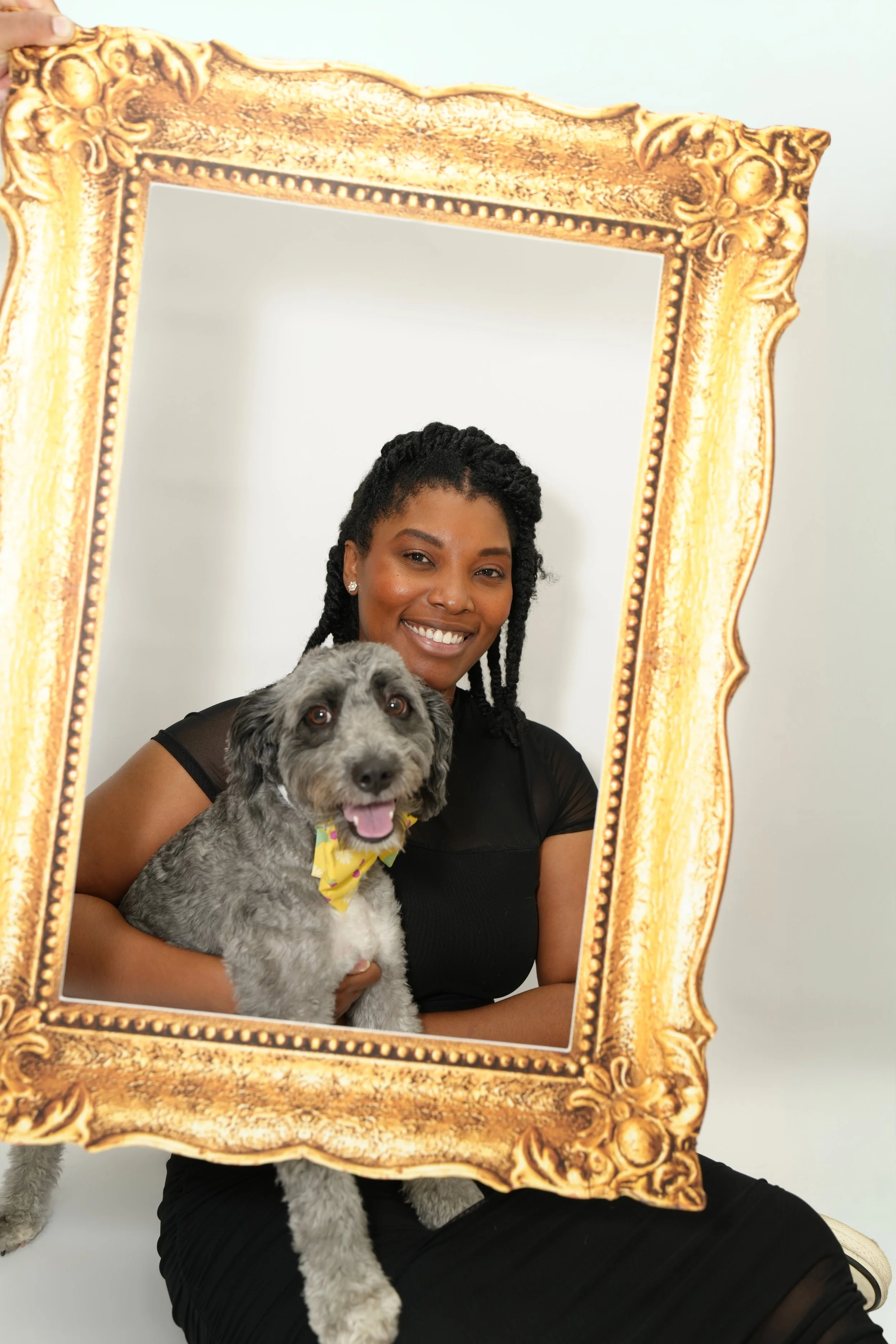 A woman with dark hair styled in twist braids is smiling while holding a gray and white dog with a yellow bow tie in front of a large ornate gold picture frame. The woman is sitting against a plain white background.