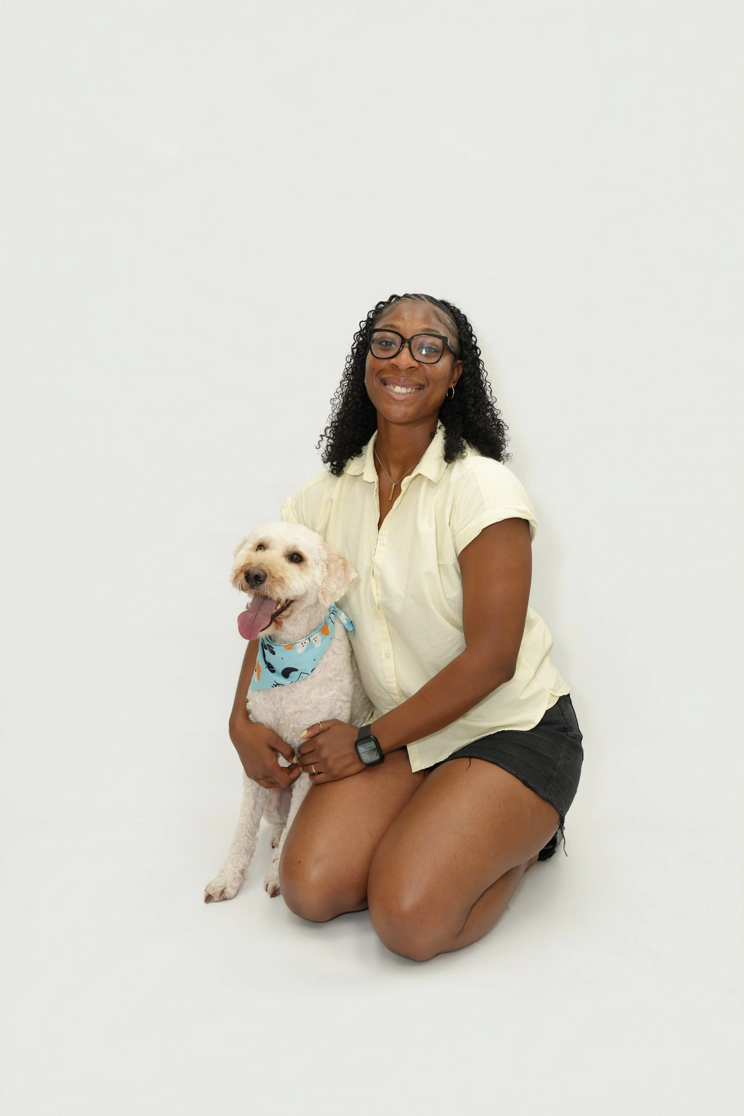 A woman with curly hair, glasses, and a yellow shirt, kneeling on one leg and holding a white dog with a blue bandana in front of a plain white wall.