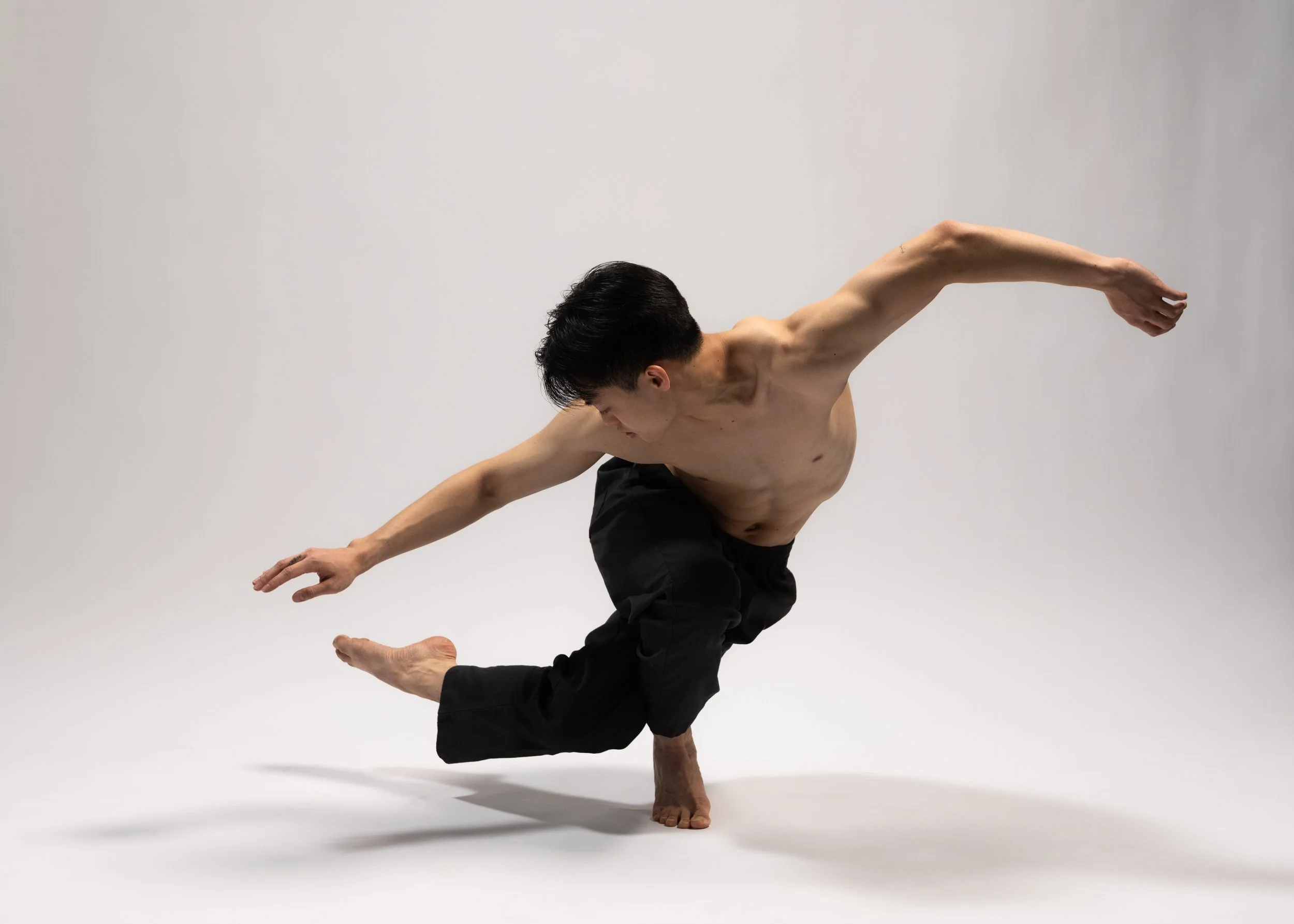 A shirtless man in black pants poses in a balancing yoga pose on a white background.