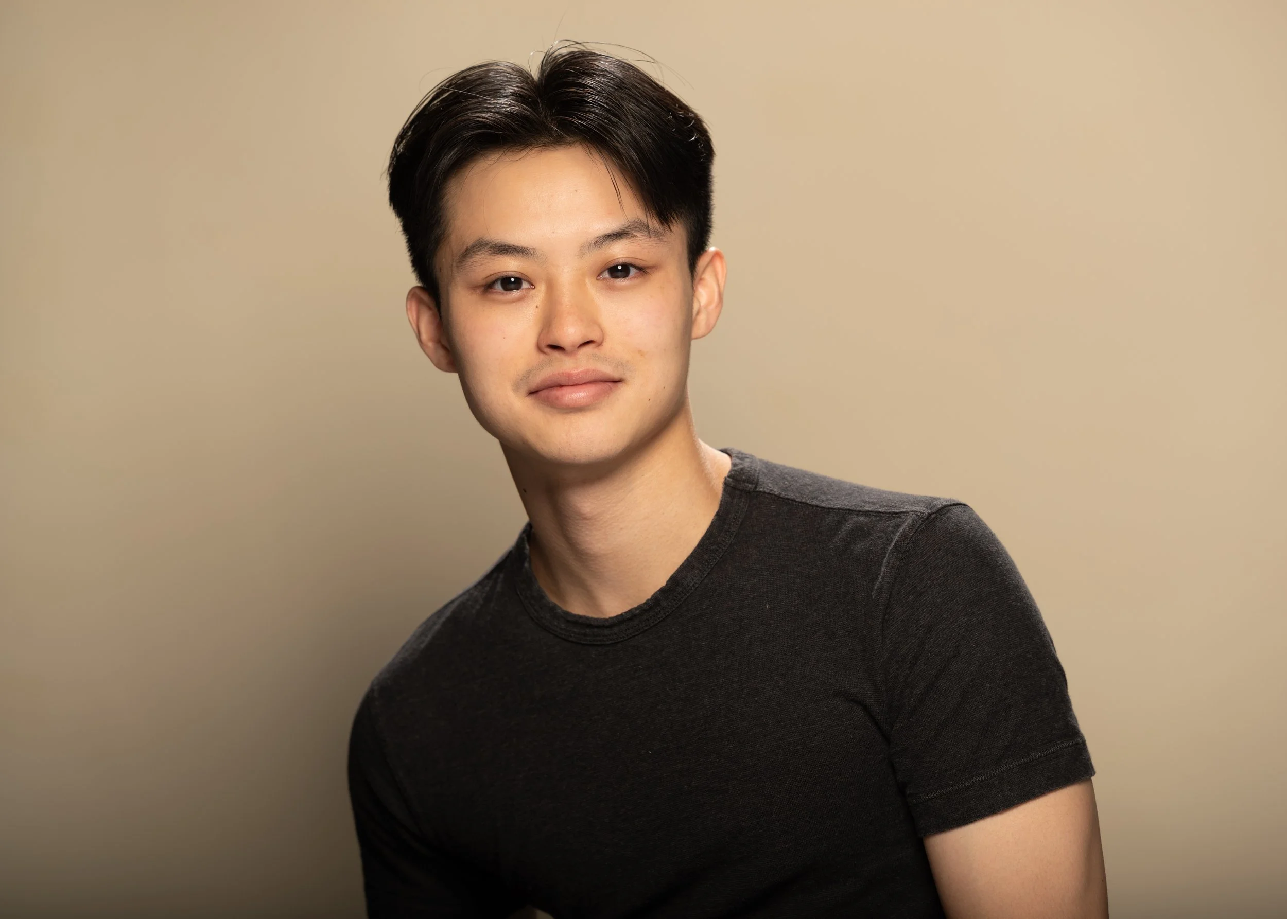 Young man with black hair wearing a black t-shirt posing against a plain beige background.