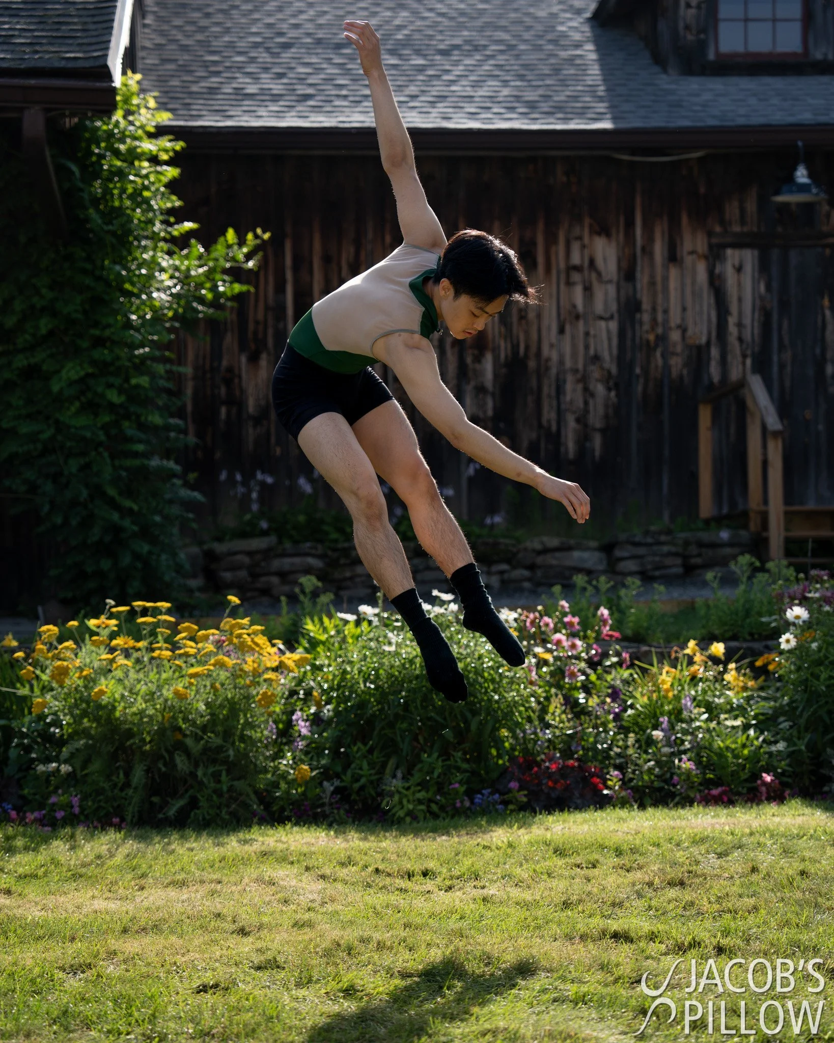 A person jumping in a backyard garden with colorful flowers, wearing a sleeveless shirt, shorts, and black socks.