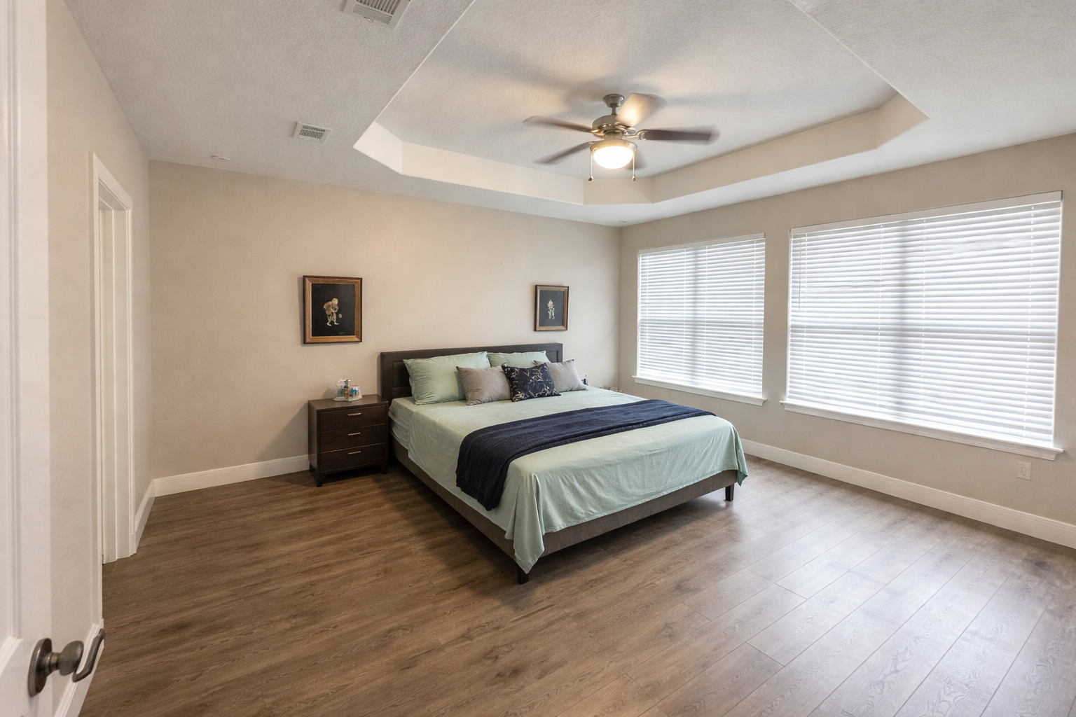 A bedroom with a large bed, nightstand, framed art, and three windows with blinds, ceiling fan, and hardwood flooring.
