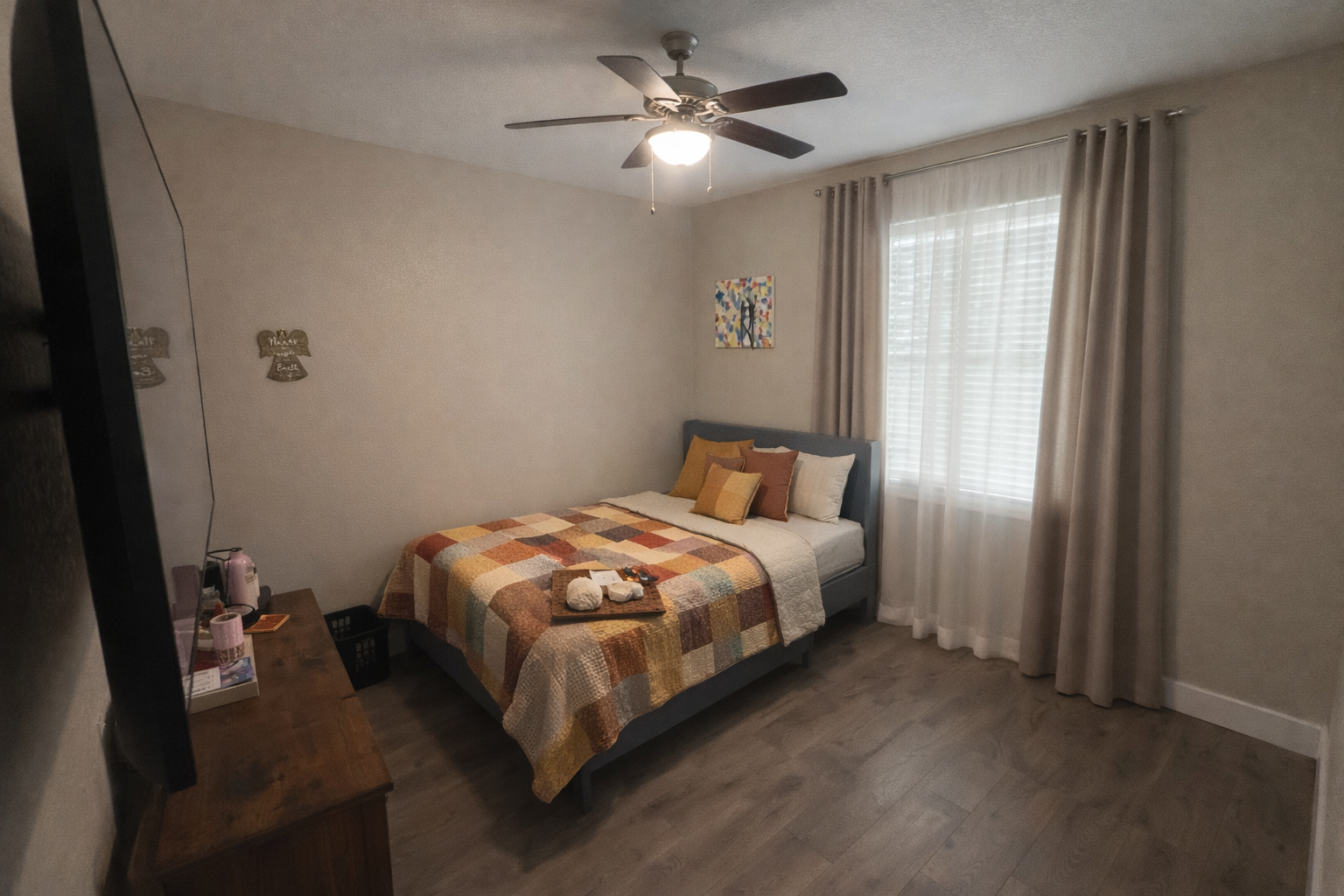 A bedroom with a bed covered by a multicolored patchwork quilt, decorated with several pillows in various shades of yellow, orange, and white. There is a small wooden dresser on the left side of the room with miscellaneous items on top. A window with white blinds and beige curtains is on the right wall, with a colorful painting hanging above the bed. A ceiling fan with a light is turned on, illuminating the room.