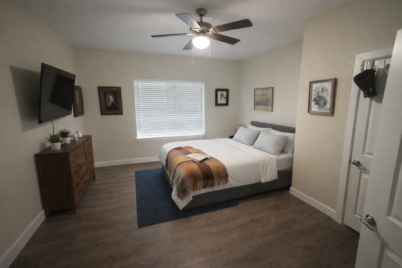 Bedroom with a bed covered in white sheets, two gray pillows, a peach and brown throw blanket, and an open book on the bed. There's a window with blinds, a wooden dresser with plants, framed artwork on the walls, a blue rug, and a ceiling fan.