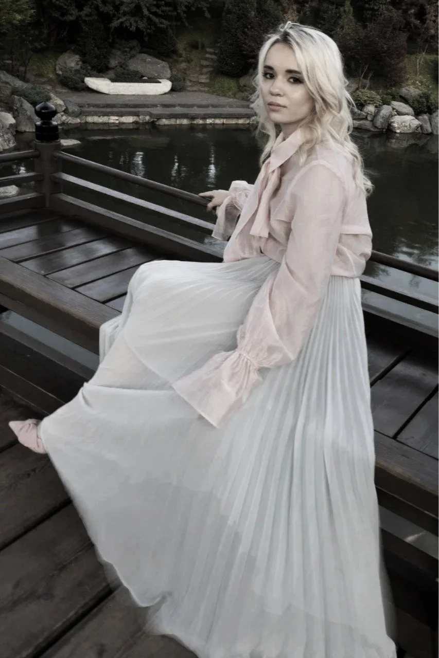 Young woman with blonde curly hair sitting on a wooden railing by a pond, wearing a light-colored pleated skirt and a pastel pink blouse, overlooking the water with trees and rocks in the background.