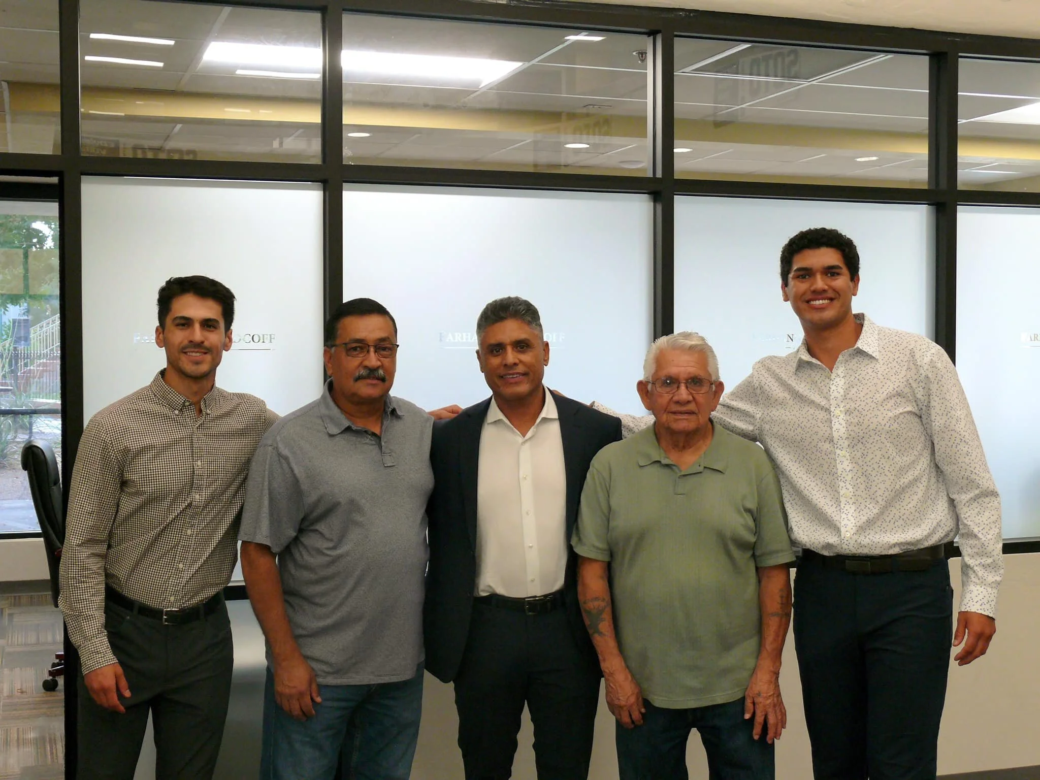 A group of five men standing together indoors, smiling at the camera with a modern office window behind them.