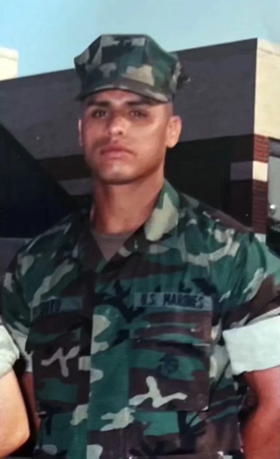 A soldier in military camouflage uniform and cap, standing outdoors in front of a building with a dark overhang and brick accents.