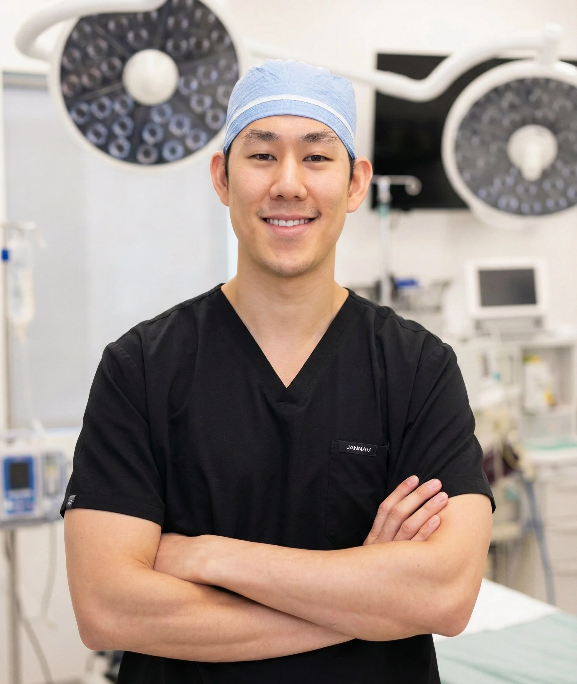 A young male healthcare professional standing with arms crossed in an operating room, wearing black scrubs and a light blue surgical cap, with surgical lights in the background.