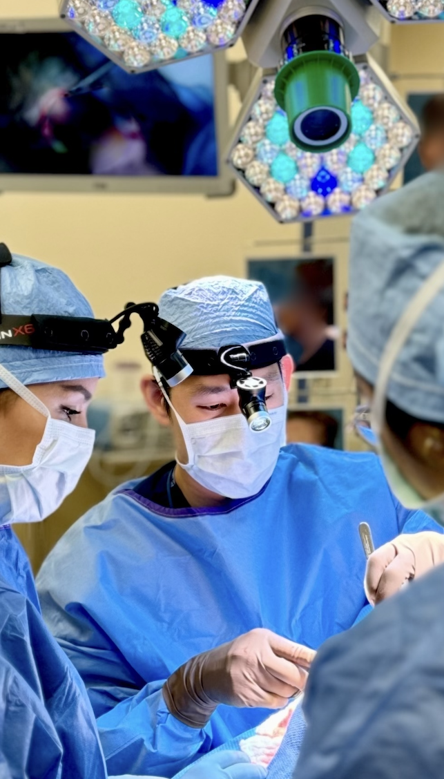 Surgeons wearing surgical masks, caps, and scrubs performing an operation in an operating room, with bright surgical lights overhead.