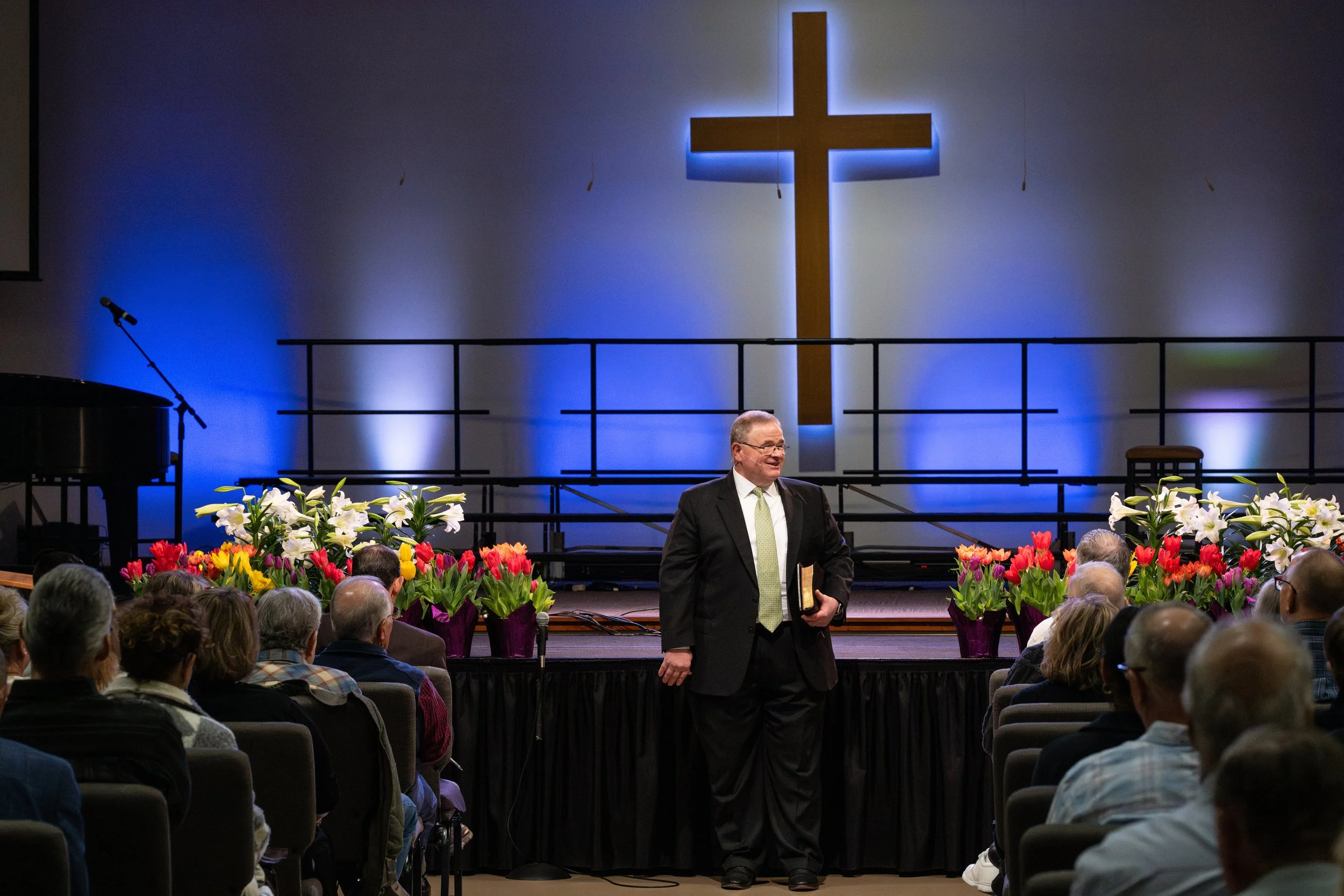 A man in a black suit and glasses stands on stage holding a book, speaking to an audience in a church or auditorium with a large wooden cross illuminated by blue lights in the background. The stage is decorated with white lilies and colorful tulips in purple pots.