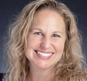 Close-up portrait of a woman with long, wavy blonde hair smiling against a dark background.
