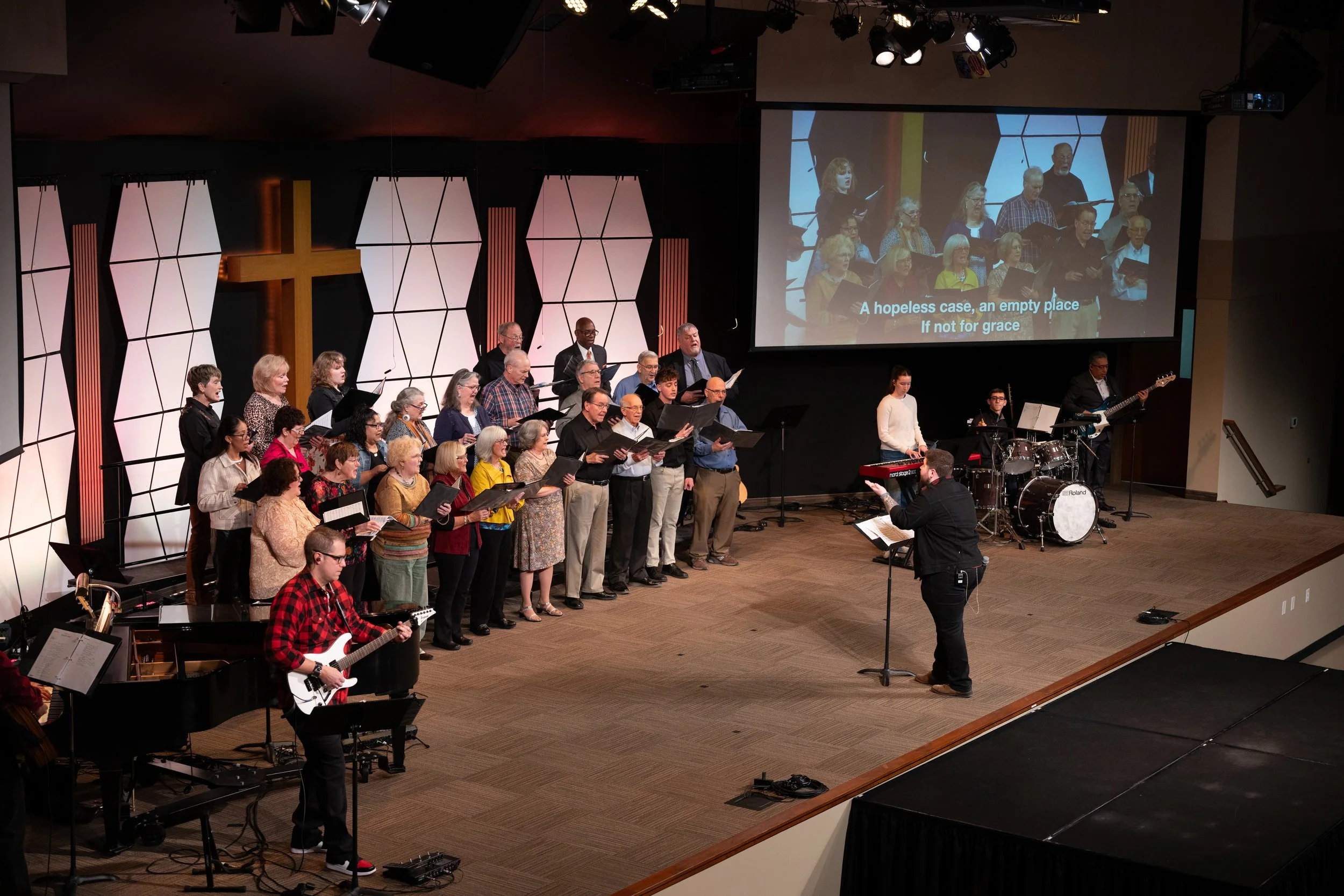 A choir performing on stage during a church service with a large cross in the background, a church service projected on a screen, and musicians playing instruments.
