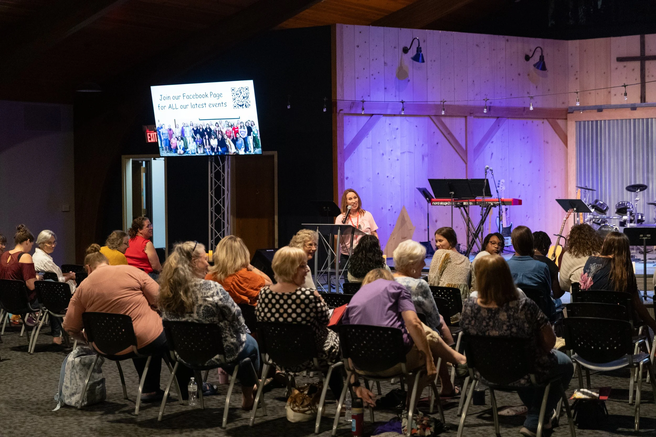 A woman speaking at a podium in front of a seated audience inside a venue with a wooden wall backdrop and musical instruments on stage. A large screen displays an invitation to join their Facebook page for latest events.