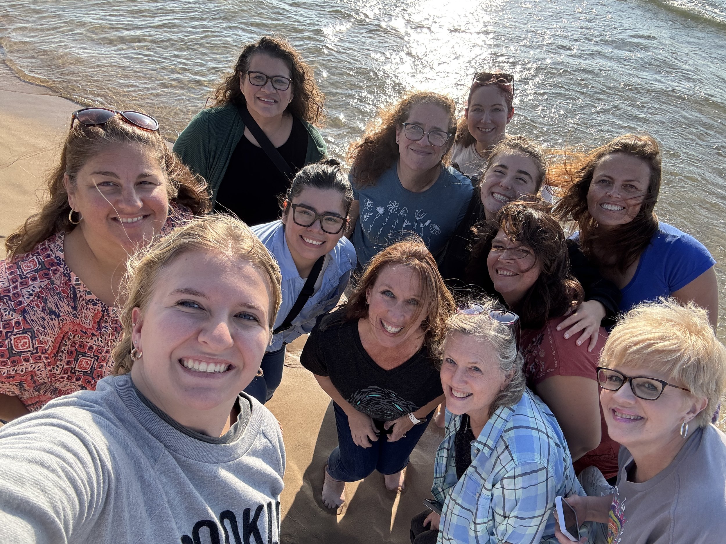 Group of women smiling for a selfie on the beach, with ocean water in the background.
