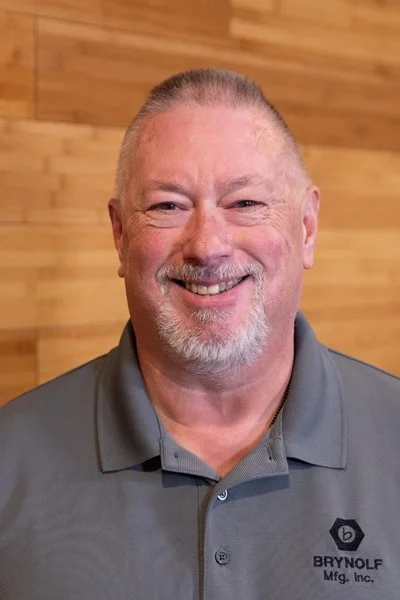 Man with gray hair and beard smiling, wearing a gray polo shirt with 'Brynoif Mfg. Inc.' logo, standing in front of a wooden wall.