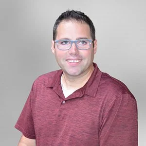 A man with short dark hair, glasses, and a light beard, smiling and wearing a maroon polo shirt against a plain gray background.
