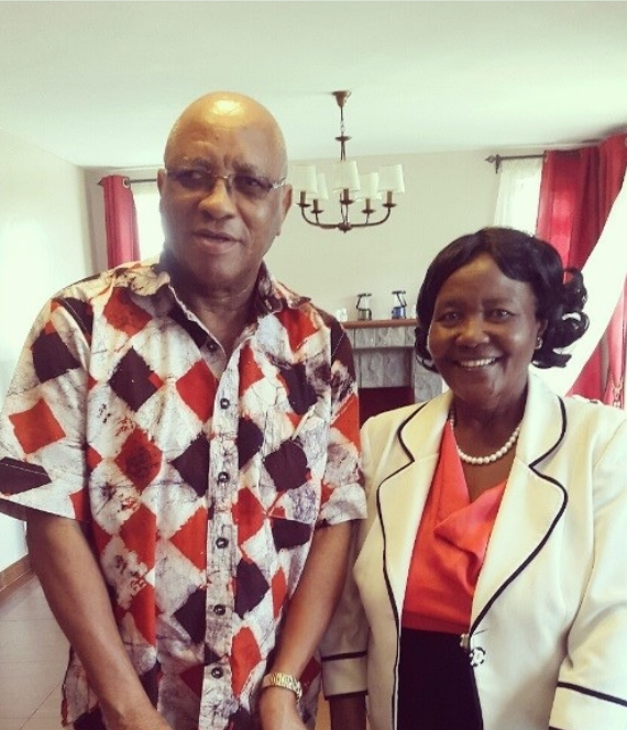 A man and woman standing indoors, smiling for the camera. The man is wearing a patterned short-sleeve shirt, glasses, and a watch. The woman is wearing a white blazer with black trim, an orange top, and a pearl necklace.