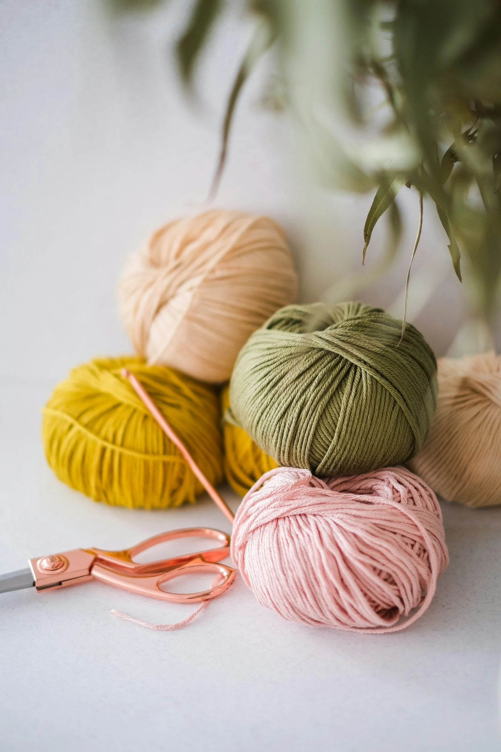 Balls of yarn in pink, green, yellow, and cream colors with a pair of pink scissors on a white surface.
