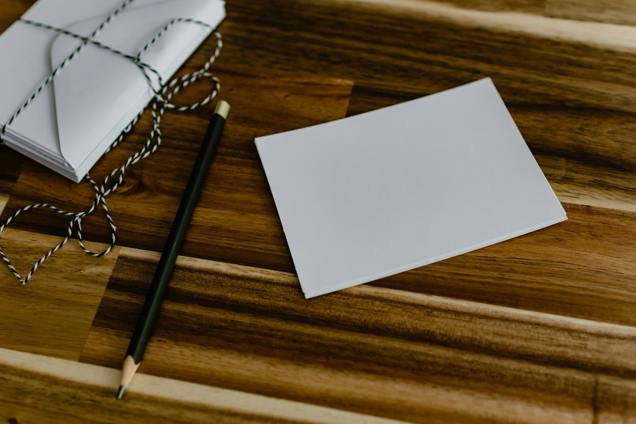 A wooden desk with a stack of white envelopes tied with black and white string, a black pen with a gold tip, a blank white card, and a blank piece of paper.