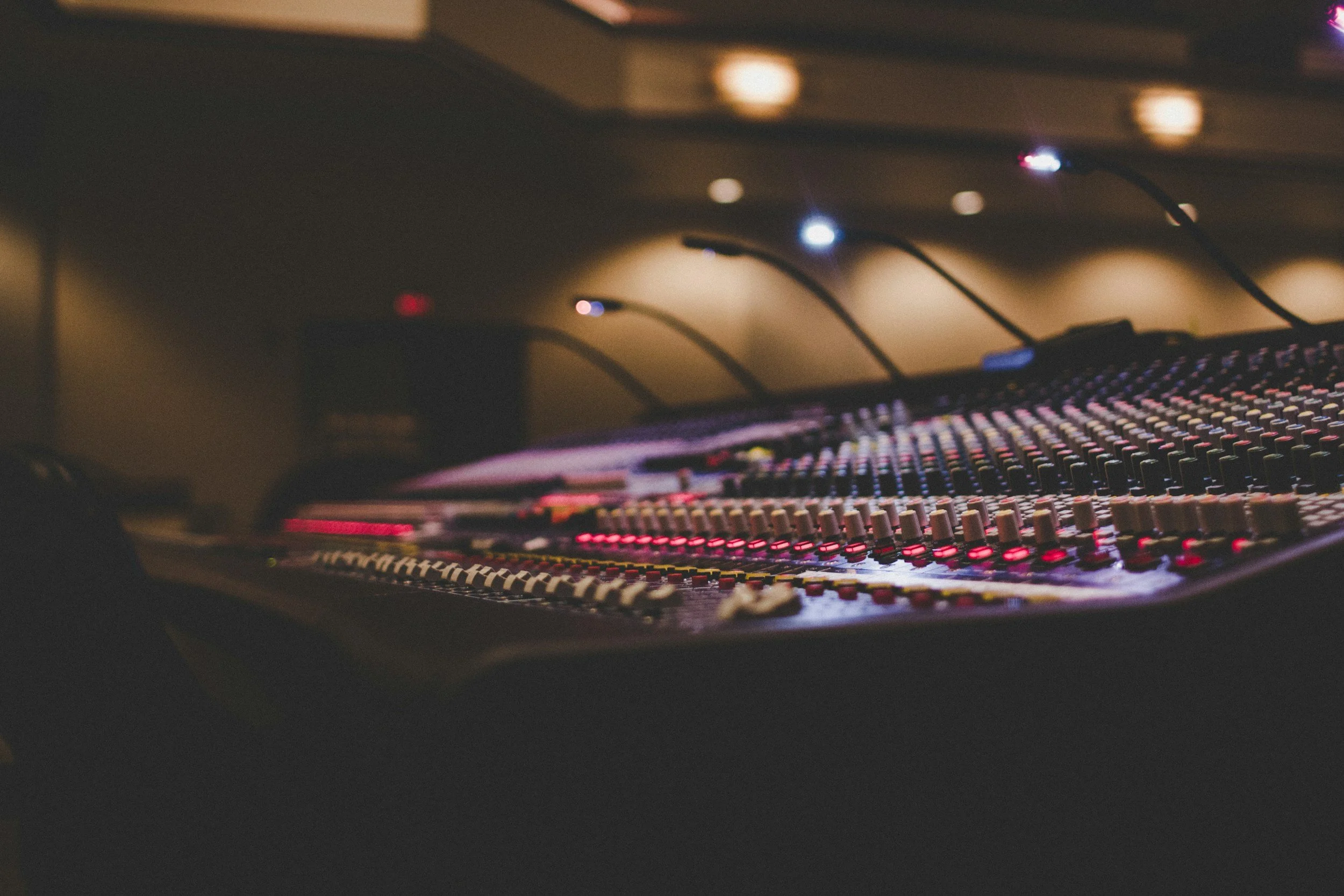 Close-up of a professional audio mixing console with various knobs and faders, under dim ambient lighting with small blue and yellow lights in the background.