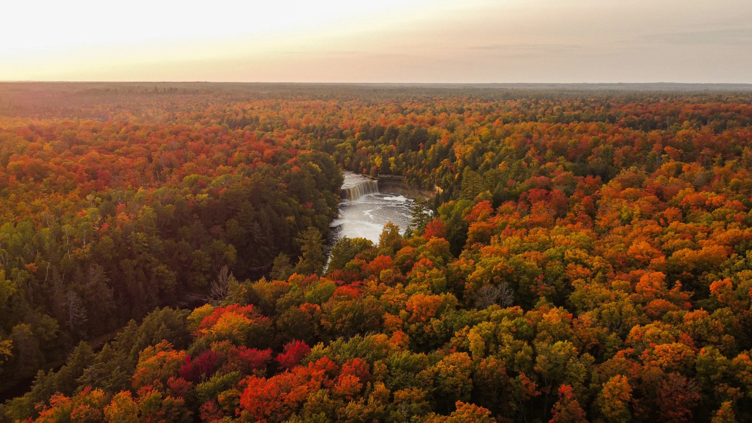 An aerial view of a river with a waterfall surrounded by a dense forest in fall, showing trees with red, orange, and green leaves.