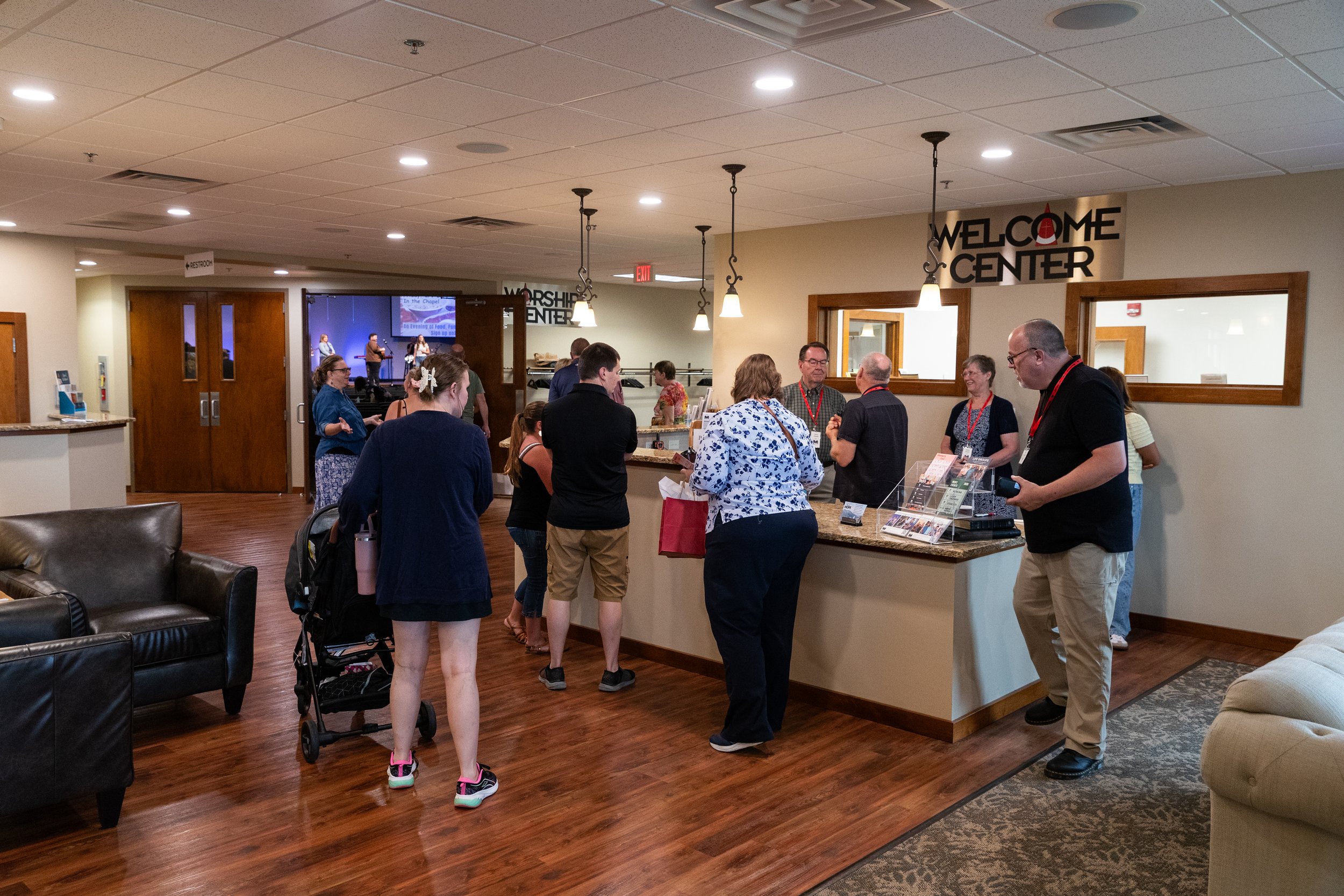 People standing in line and talking at a reception desk in a church lobby, with a sign reading 'Welcome Center' above. There are church members and visitors, some wearing name tags, and a worship band visible on a stage in the background.