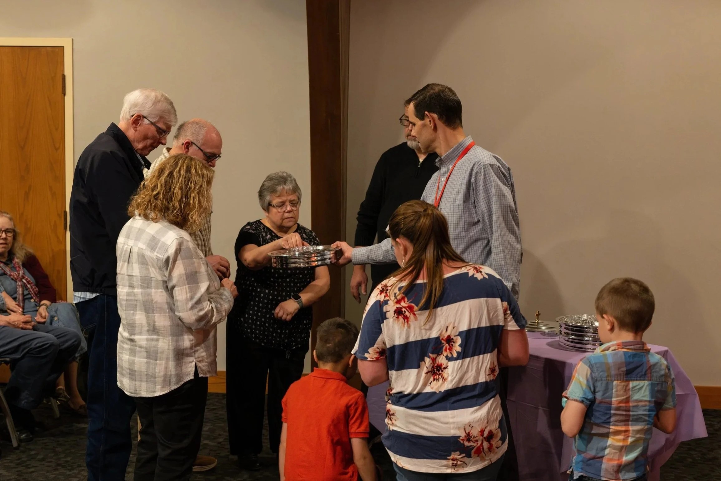 A group of people gathered around a table with metal plates, with some individuals standing and others seated, inside a room with beige walls and wooden accents.