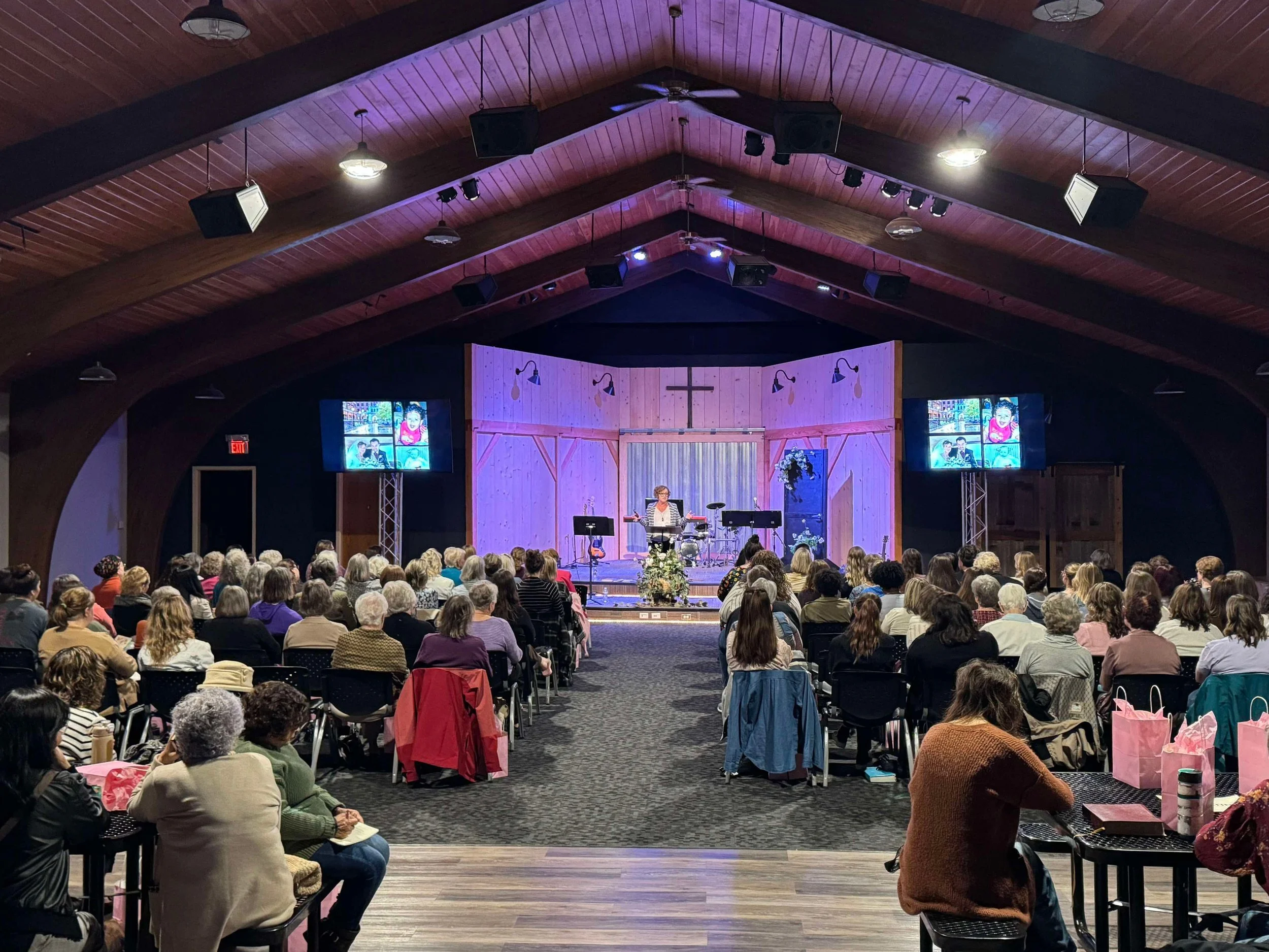 A church or auditorium with a wooden ceiling and a stage at the front. The stage has a cross, floral decorations, a woman speaking at a podium, and two large screens displaying a presentation. The audience is comprised mostly of women seated facing the stage.