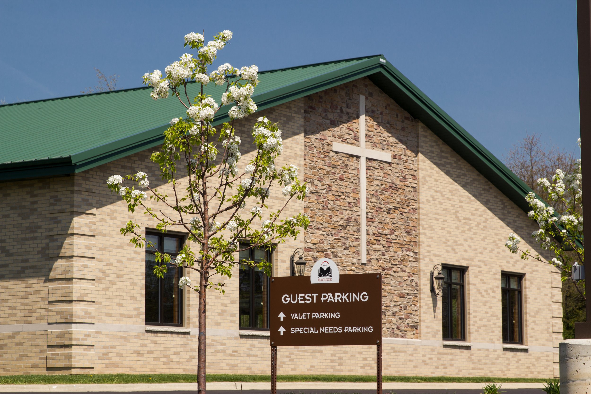 A church building with a green roof, brick and stone walls, and a white cross affixed to the front. In front of the building, there is a sign indicating guest parking, valet parking, and special needs parking. A flowering tree with white blossoms is positioned to the left of the sign.