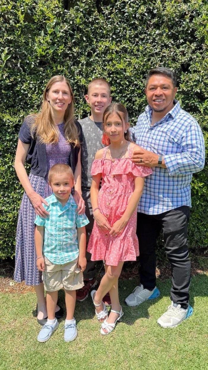 A family of five posing outdoors in front of a green hedge. The woman has long blonde hair and is wearing a purple top with a patterned skirt. The man has short dark hair and is wearing a blue checkered shirt with black pants. The boy has short hair and is wearing a gray patterned shirt with black pants. The girl has long hair in pigtails and is wearing a pink dress with ruffles. The young boy has short hair and is wearing a turquoise plaid shirt with beige shorts.