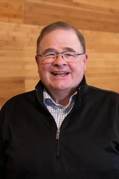 A man smiling, wearing glasses, a checkered shirt, and a black zip-up jacket, standing against a wooden wall.