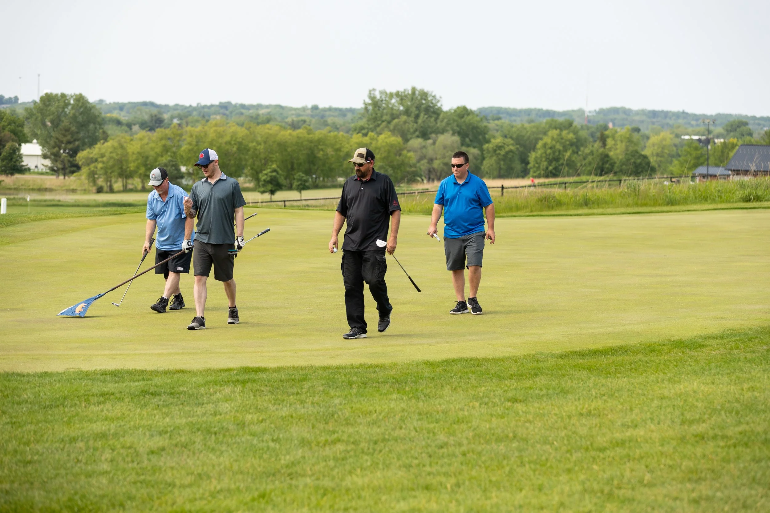 Four men walking on a golf course, with one using a rake to smooth the green, surrounded by greenery and houses in the background on a cloudy day.