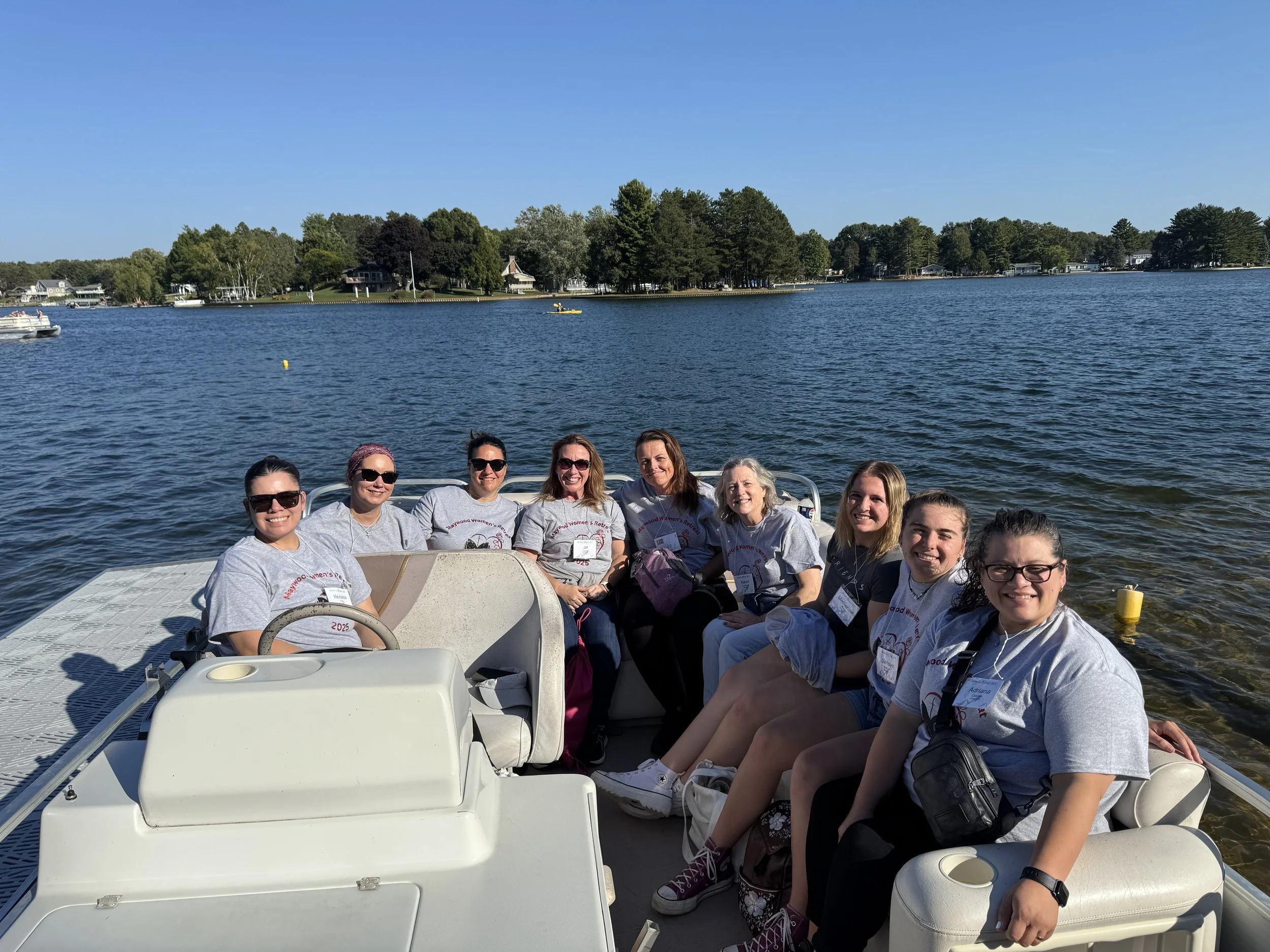 A group of nine women sitting on a boat by a lake, all wearing matching gray t-shirts and smiling at the camera, with trees and houses visible across the water.