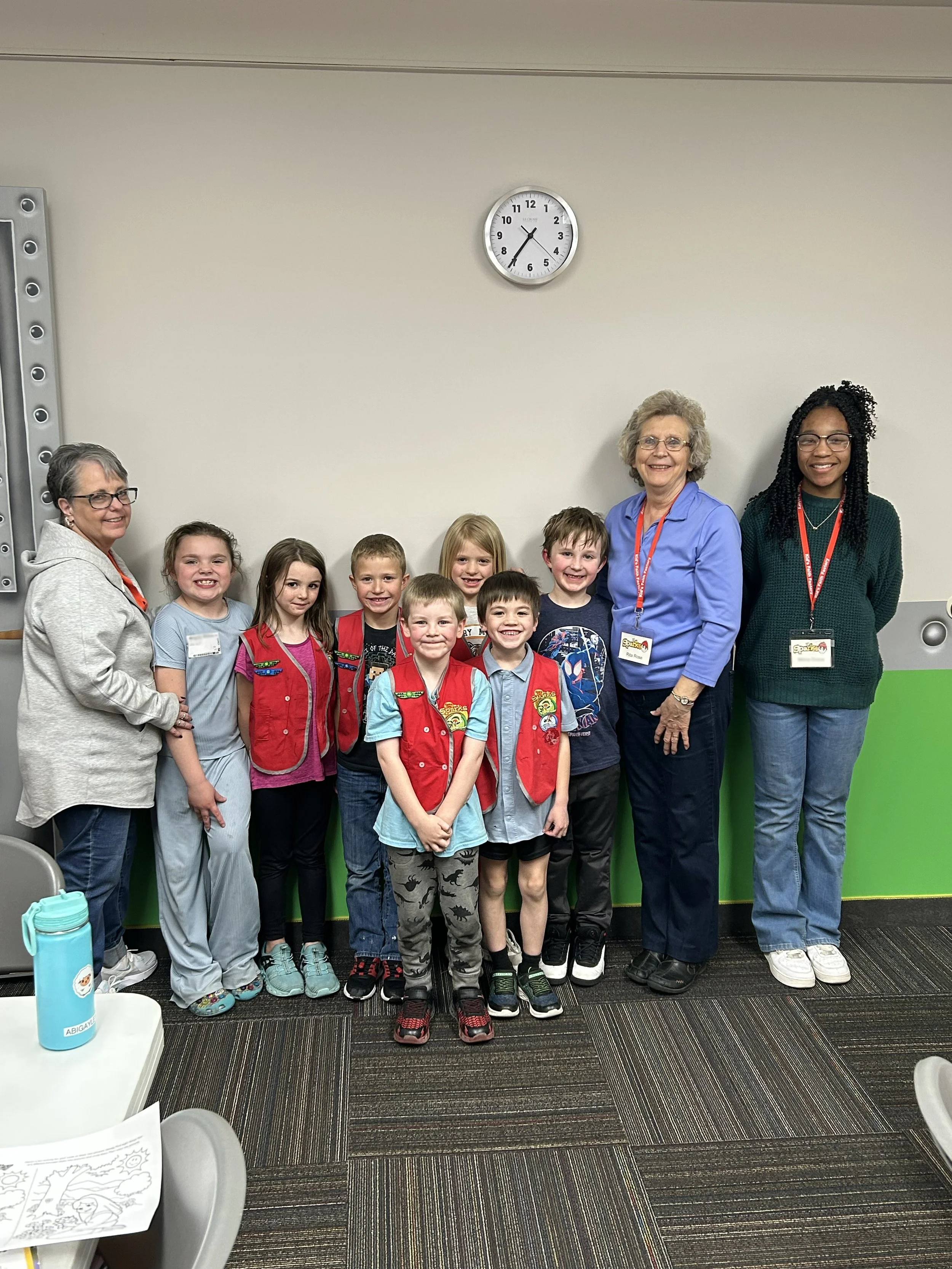 A group of children and adults posing for a photo in a classroom or community center, with a plain wall and a clock showing 1:25 in the background.