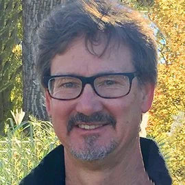 A man with glasses and dark, curly hair smiling outdoors in front of a tree with autumn leaves.