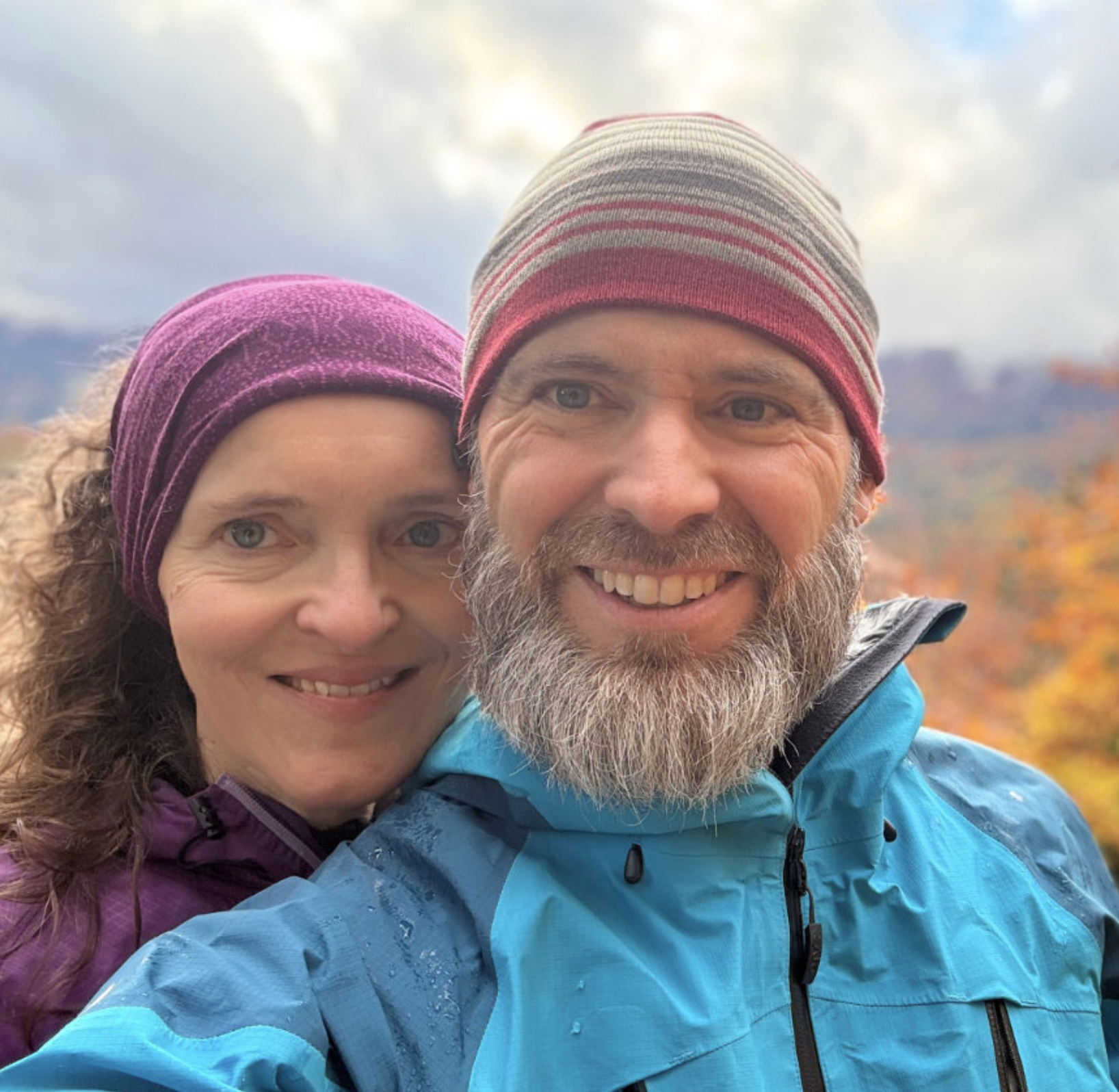 Close-up of a smiling couple outdoors in fall, wearing colorful jackets and beanies, with autumn trees and mountains in the background.
