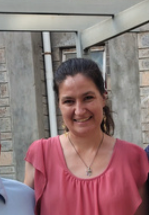 Smiling woman with dark hair in a ponytail, wearing a pink top and a necklace, standing outdoors near a concrete wall with industrial pipes.