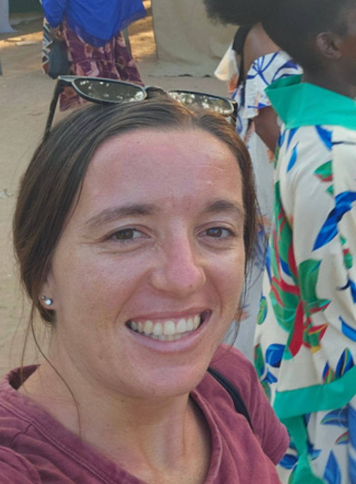 Close-up of a smiling woman with sunglasses on her head, standing outdoors with other people around.