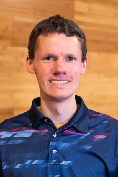 A man with short brown hair smiling at the camera, wearing a blue patterned collared shirt, standing in front of a wooden wall background.