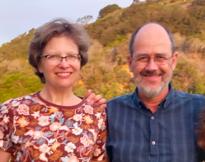 A smiling woman with short gray hair and glasses, and a smiling man with glasses and a beard, standing outdoors in front of a hillside with trees and foliage.