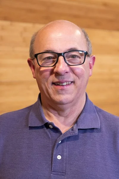 A smiling middle-aged man with glasses, wearing a purple polo shirt, standing in front of a wooden wall.