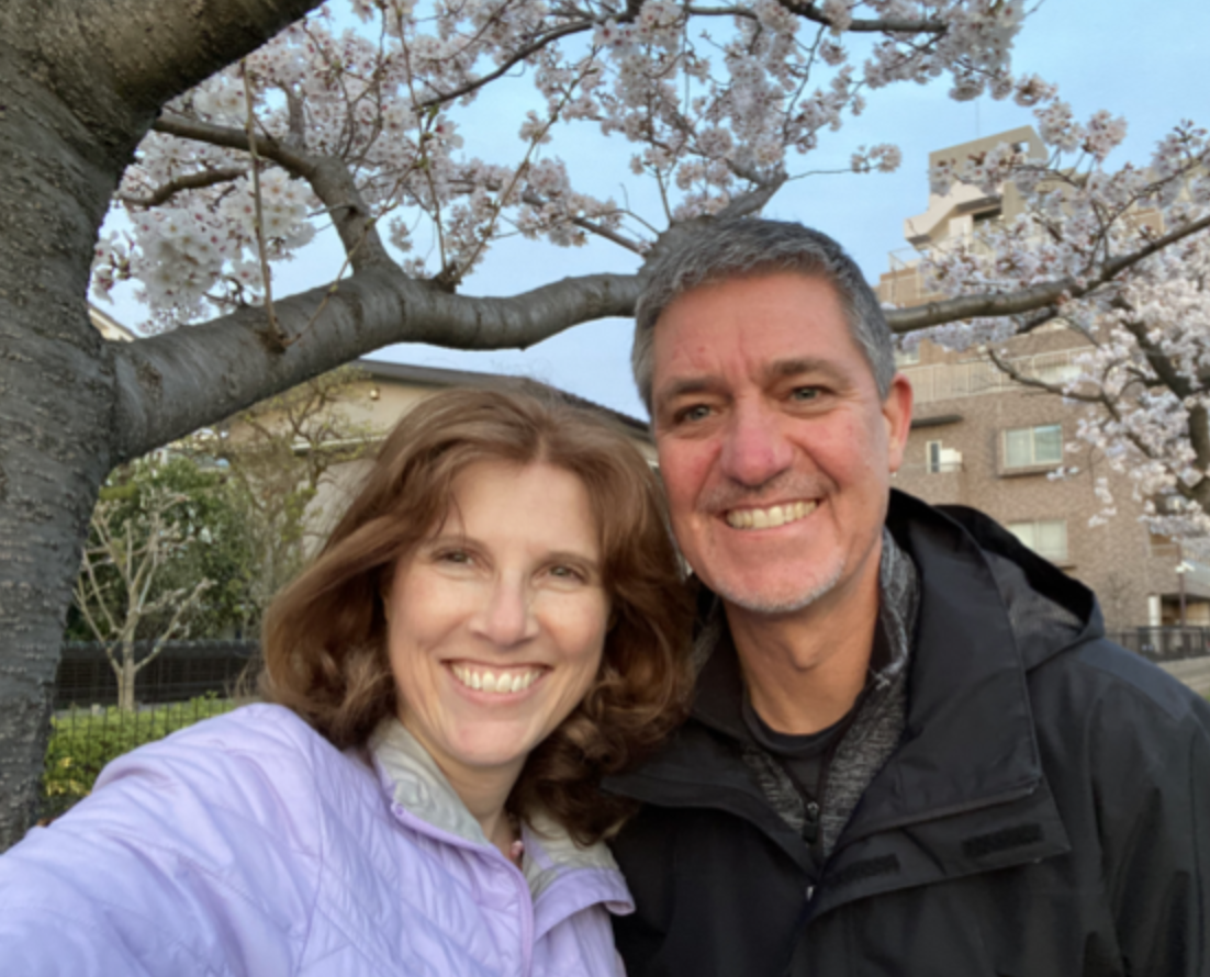 A smiling couple taking a selfie outdoors under a cherry blossom tree in bloom with a brick building in the background.