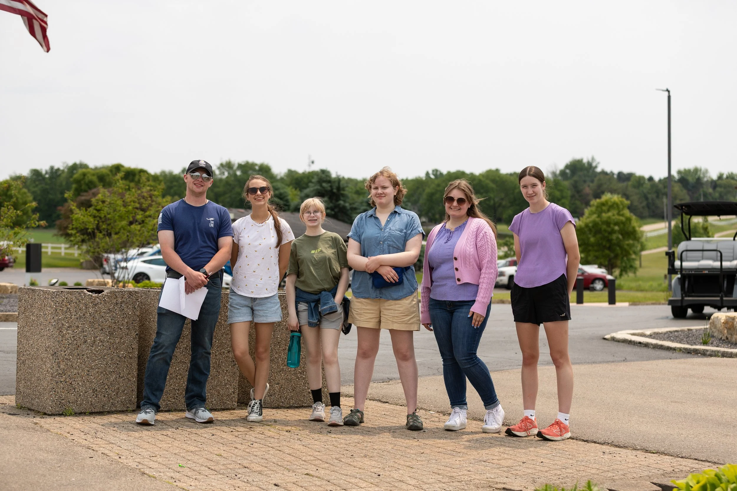 A group of seven people standing outdoors on a paved area, with a background of trees, parked cars, and a golf cart, during daytime.
