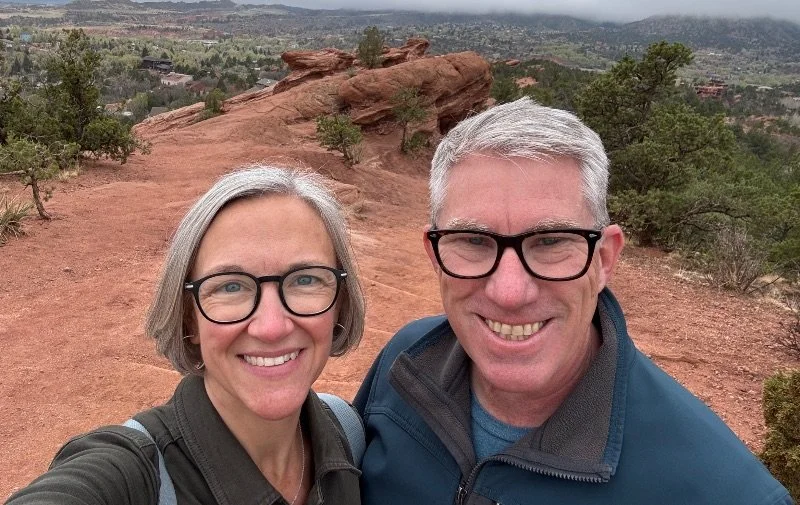 A smiling middle-aged couple wearing glasses, standing on a red rocky trail with trees and distant landscape in the background.