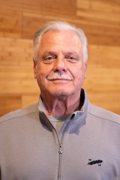 An older man with gray hair and a mustache smiling at the camera, wearing a light gray zip-up jacket with a small logo, standing in front of a wooden wall.