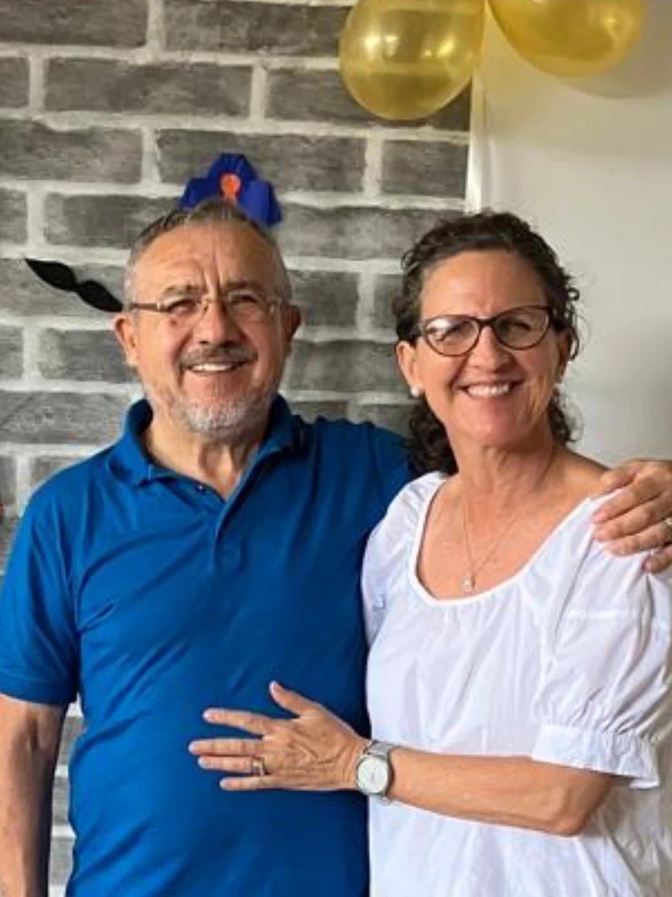 A smiling older man and woman standing close together, the woman with her hand on the man's chest, in front of a gray brick wall and gold balloons.