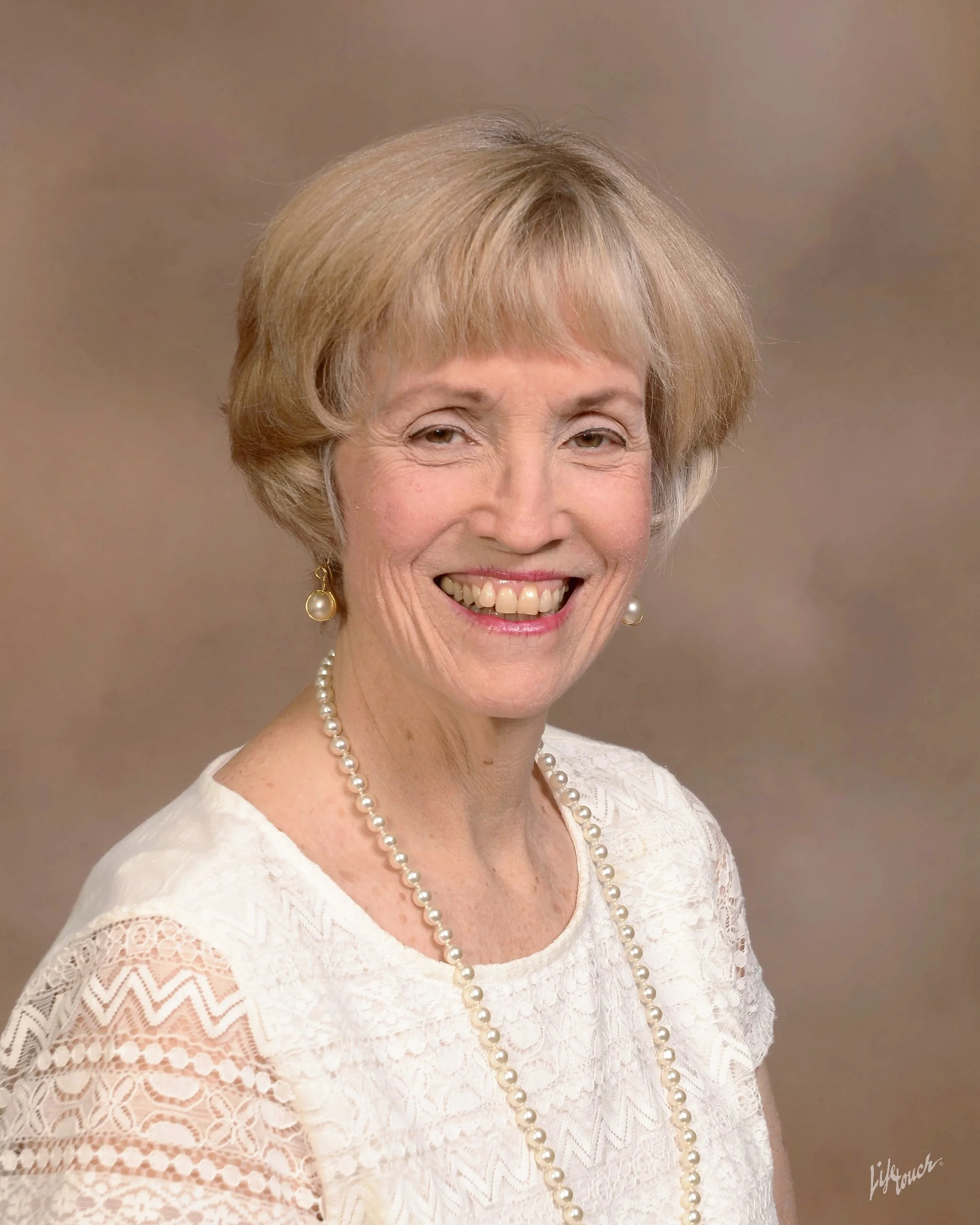 Portrait of an elderly woman with short blonde hair, wearing a white lace top, pearl necklace, and pearl earrings, smiling against a neutral background.
