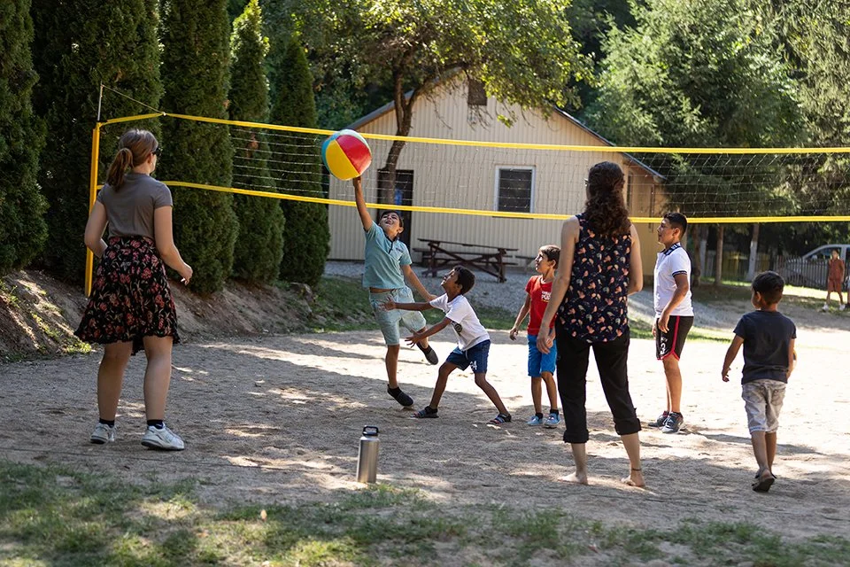 Group of children and adults playing volleyball outdoors on a sunny day, with trees and a house in the background.