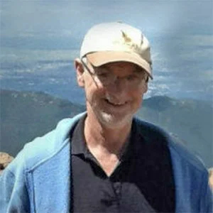 A smiling man wearing a white cap, glasses, a blue jacket, and a black shirt, outdoors with mountains in the background.